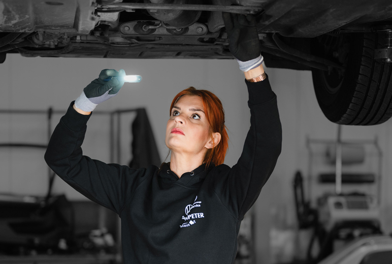 Die Kfz-Mechatronikerin Anne Althaus-Braun inspiziert den Unterbodens eines aufgebockten Autos mit einer Leuchte.