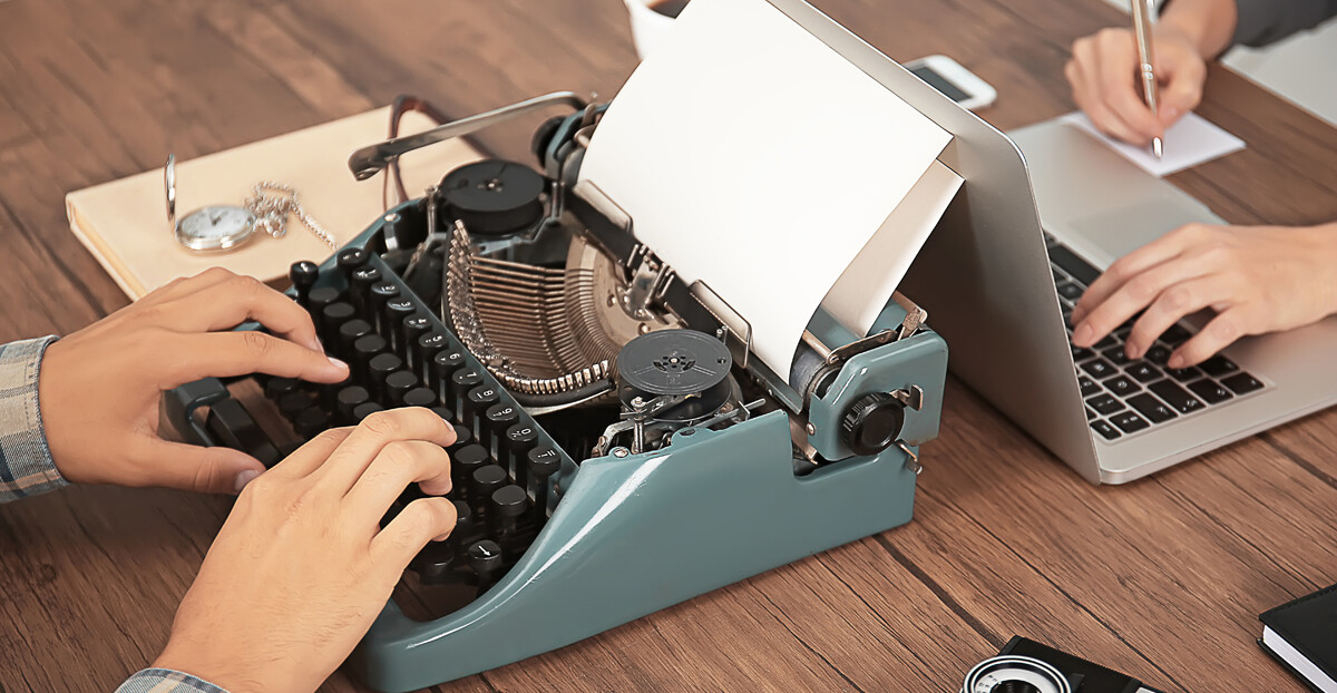 Detailaufnahme von Arbeitsplatz: Tisch mit Schreibmaschine und Laptop gegenüber mit tippenden Händen
