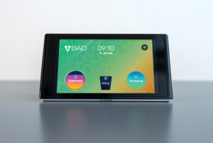 Ein schwarzes, rechteckiges Displaygerät steht auf einer glatten Oberfläche. Der Bildschirm zeigt eine Benutzeroberfläche mit dem Logo „BAD“ oben links, der Uhrzeit „09:10“ und dem Datum „Fr., 20. Mai“. Darunter befinden sich zwei runde Symbole mit den Bezeichnungen „Ergonomie“ (links, in Lila und Orange) und „Bewegung“ (rechts, in Blau). In der Mitte wird eine Wassertrinkanzeige mit „0.0/1.5L“ dargestellt. Der Hintergrund des Displays ist grün-gelb mit einem dezenten Muster.