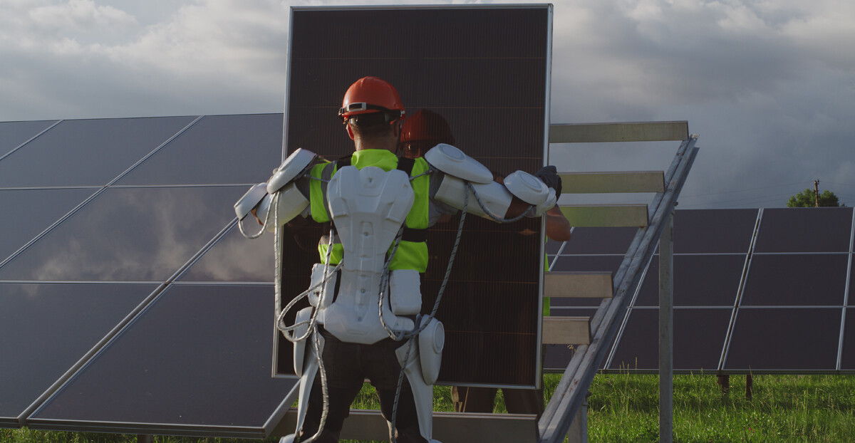 Person mit Exoskelett auf einer Wiese, die eine Solarpanele hält, dahinter ein schräges Gestell zur Anbringung. Daneben und davor stehen weitere schon montierte, in etwa 90 Grad aufgerichtete Solarpanelen auf Gras.