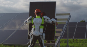 Person mit Exoskelett auf einer Wiese, die eine Solarpanele hält, dahinter ein schräges Gestell zur Anbringung. Daneben und davor stehen weitere schon montierte, in etwa 90 Grad aufgerichtete Solarpanelen auf Gras.