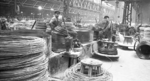 Ein schwarz-weiß Bild einer Industrie-Halle. Zu sehen sind drei Männer die in die Kamera gucken und haufenweise Draht.
