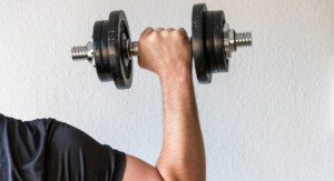 Nahaufnahme eines muskulösen Männerarms, der eine schwarze Kurzhantel mit festem Griff hält. Der Bildausschnitt fokussiert sich auf den Arm und die Hantel, während der Hintergrund eine schlichte weiße Wand ist.
