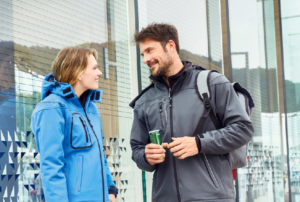 Ein Mann in schwarzer Softshell Jacke und eine Frau in blauer Softshell Jacke stehen draußen vor einer Fensterfront und unterhalten sich. Der Mann trägt einen grauen Rucksack und hält eine grüne Getränkedose.