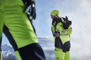 Eine Frau trägt leuchtend gelbe und schwarze Schutzkleidung mit reflektierenden Streifen, inklusive Mütze und Jacke, und steht vor einer verschneiten Berglandschaft. Eine zweite Person ist teilweise im Vordergrund sichtbar.