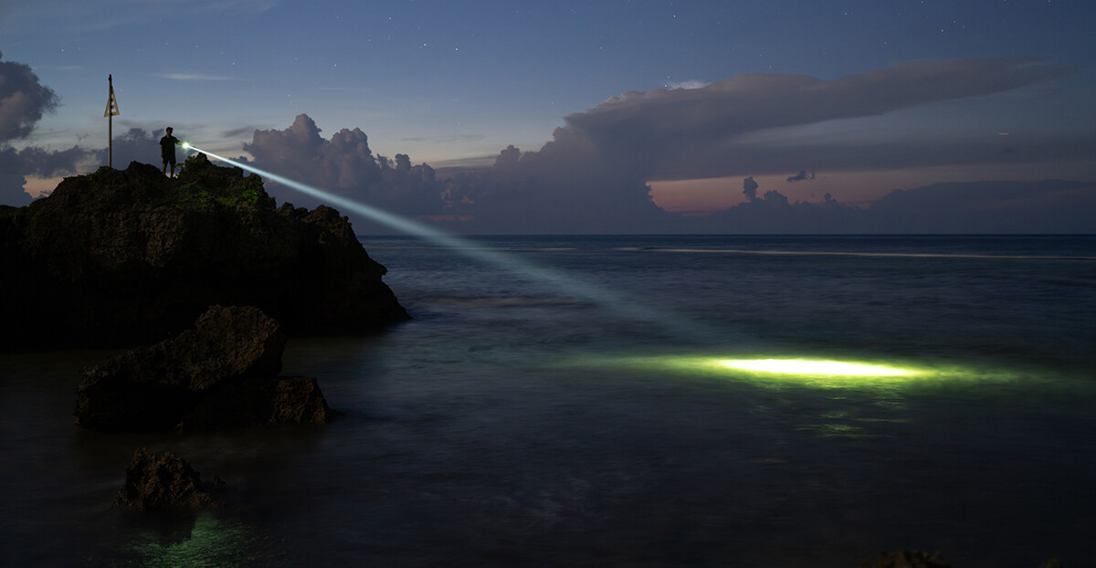 Eine Person steht bei Dämmerung oder Nacht auf einem Felsen am Meer und leuchtet mit einer leistungsstarken Taschenlampe über das Wasser. Der Lichtstrahl ist deutlich sichtbar und trifft weit draußen auf die Wasseroberfläche. Am Himmel sind Sterne und Wolken zu erkennen.