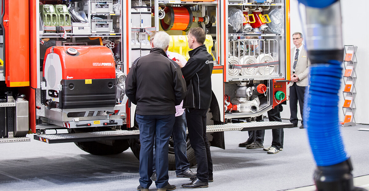 Mehrere Personen stehen vor einem geöffneten Einsatzfahrzeug der Feuerwehr, das mit verschiedenen technischen Geräten und Ausrüstungsgegenständen ausgestattet ist. Ein Fachmann erklärt den Anwesenden Details der Ausrüstung. Im Hintergrund beobachtet ein Mann im Anzug die Szene. Die Umgebung deutet auf eine Messe oder Fachausstellung hin.