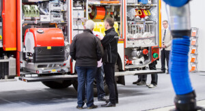 Mehrere Personen stehen vor einem geöffneten Einsatzfahrzeug der Feuerwehr, das mit verschiedenen technischen Geräten und Ausrüstungsgegenständen ausgestattet ist. Ein Fachmann erklärt den Anwesenden Details der Ausrüstung. Im Hintergrund beobachtet ein Mann im Anzug die Szene. Die Umgebung deutet auf eine Messe oder Fachausstellung hin.