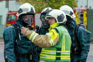 Ein Gruppenleiter der Feuerwehr, erkennbar an seiner Weste mit der Aufschrift „Abschnitt Umwelt“, gibt mehreren Feuerwehrkräften in Schutzanzügen und Atemschutzmasken Anweisungen. Im Hintergrund sind Feuerwehrfahrzeuge zu sehen. Die Szene wirkt wie eine koordinierte Einsatzbesprechung bei einem Gefahrstoffeinsatz.