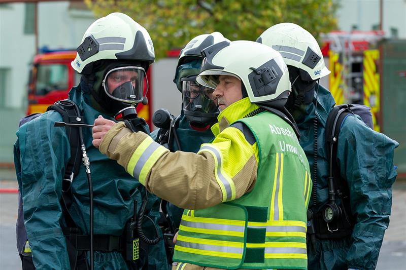 Ein Gruppenleiter der Feuerwehr, erkennbar an seiner Weste mit der Aufschrift „Abschnitt Umwelt“, gibt mehreren Feuerwehrkräften in Schutzanzügen und Atemschutzmasken Anweisungen. Im Hintergrund sind Feuerwehrfahrzeuge zu sehen. Die Szene wirkt wie eine koordinierte Einsatzbesprechung bei einem Gefahrstoffeinsatz.