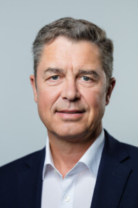 Ein Mann mit weißen Hed und schwarzem Sakko guckt in die Kamera.