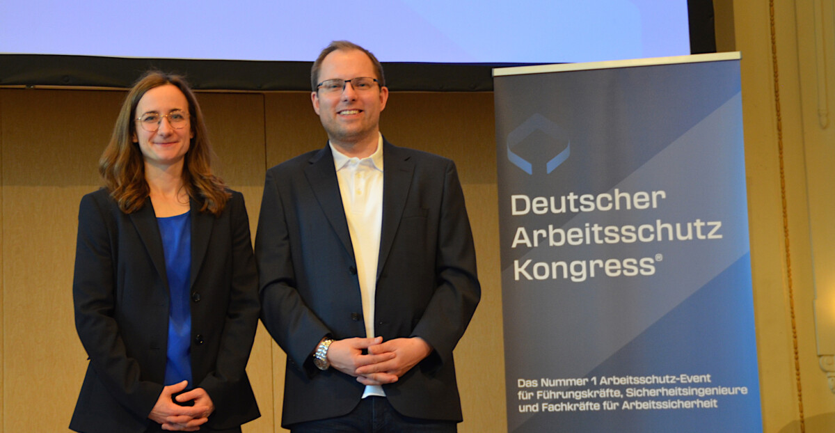 Ein Mann und eine Frau posieren vor einem Aufsteller mit der Aufschrift "Deutscher Arbeitsschutz Kongress".