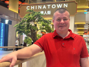 Eine Person in rotem Poloshirt steht in einem Einkaufszentrum vor einem Schild mit der Aufschrift „CHINATOWN“ und asiatischen Schriftzeichen. Im Hintergrund sind ein Baum und mehrere Menschen zu sehen.