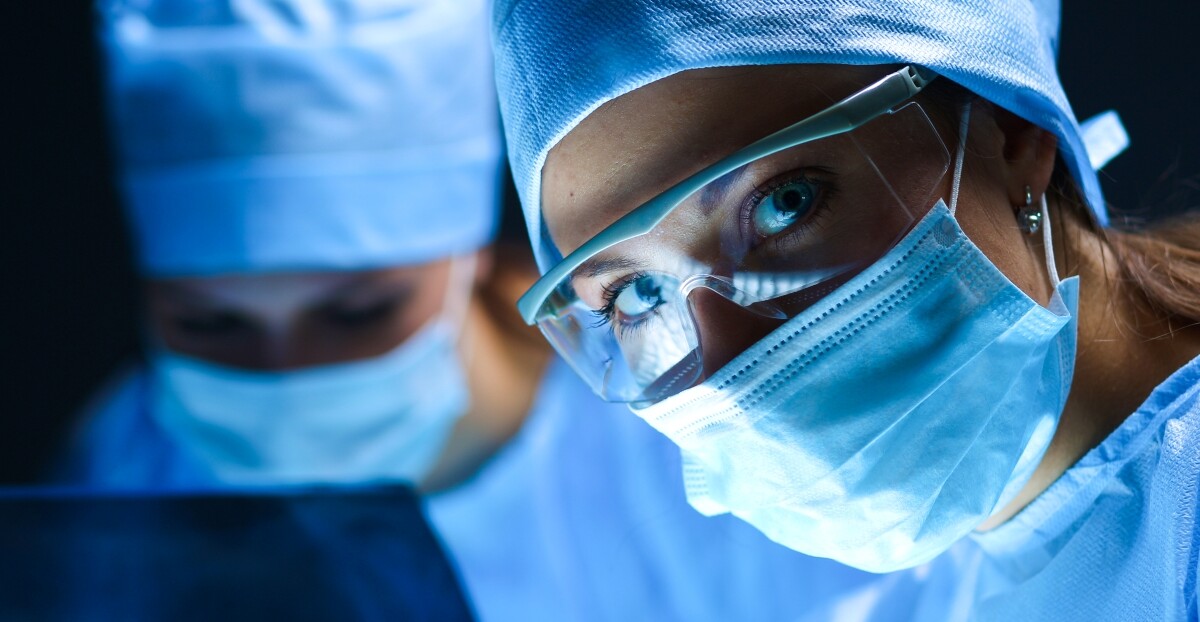Zwei Personen in OP-Kleidung und mit Mundschutz, von denen die Frau im Vordergrund eine Schutzbrille trägt und direkt in die Kamera blickt.