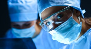 Zwei Personen in OP-Kleidung und mit Mundschutz, von denen die Frau im Vordergrund eine Schutzbrille trägt und direkt in die Kamera blickt.