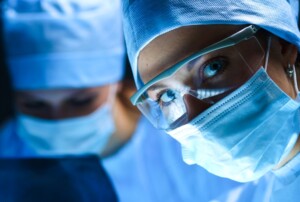 Zwei Chirurginnen in OP-Kleidung und Mundschutz arbeiten konzentriert in einem Operationssaal. Die Frau im Vordergrund trägt zusätzlich eine Schutzbrille und blickt direkt in die Kamera.