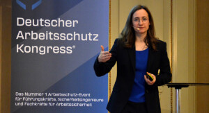 Frau steht neben einem Roll-Up mit der Aufschrift „Deutscher Arbeitsschutz Kongress“ und spricht.