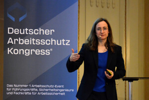Frau steht neben einem Roll-Up mit der Aufschrift „Deutscher Arbeitsschutz Kongress“ und spricht.