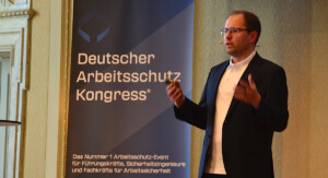 Mann steht neben einem Roll-Up mit der Aufschrift „Deutscher Arbeitsschutz Kongress“ und spricht.