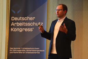 Mann steht neben einem Roll-Up mit der Aufschrift „Deutscher Arbeitsschutz Kongress“ und spricht.