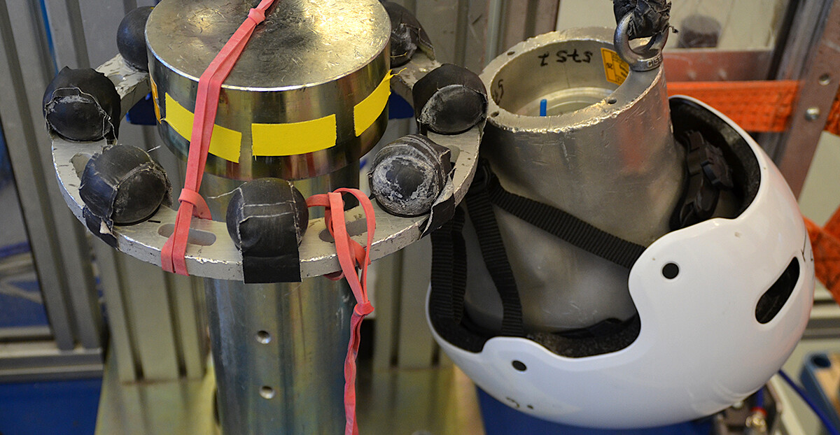 Ein Prüfstands zur Helmsicherheit. Links ist ein zylindrischer Metallpfosten mit schwarzen Sensoren und roten Gummibändern zu sehen. Rechts daneben hängt ein weißer Schutzhelm an einem Aluminiumkopfmodell, das an einem Seil befestigt ist. Im Hintergrund sind Metallrahmen und orangefarbene Sicherungsbänder sichtbar.