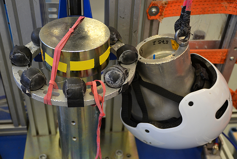 Ein Prüfstands zur Helmsicherheit. Links ist ein zylindrischer Metallpfosten mit schwarzen Sensoren und roten Gummibändern zu sehen. Rechts daneben hängt ein weißer Schutzhelm an einem Aluminiumkopfmodell, das an einem Seil befestigt ist. Im Hintergrund sind Metallrahmen und orangefarbene Sicherungsbänder sichtbar.