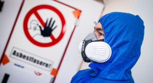 Eine Person im blauen Einwegoverall trägt eine Halbmaske mit Partikelfiltern und steht vor einer Tür mit durchgestrichenem Handsymbol und dem Warnhinweis „Asbestfasern!“.