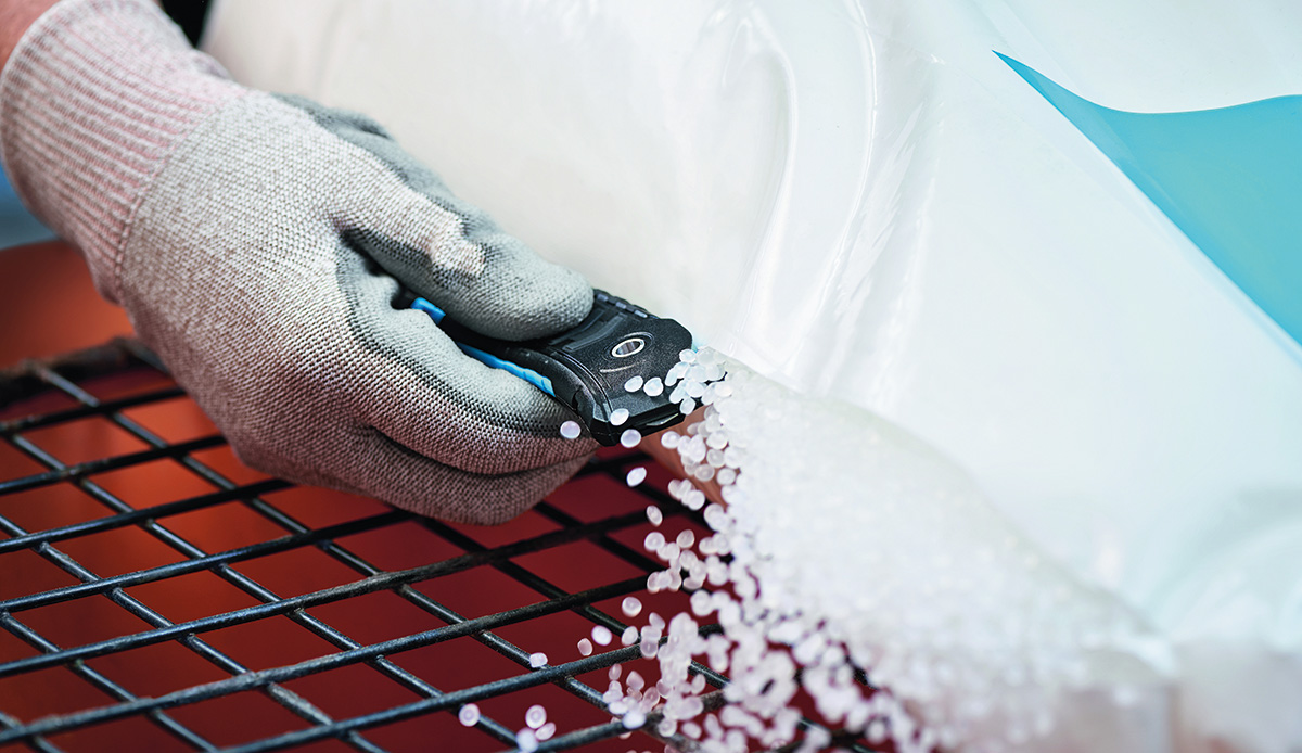 Eine behandschuhte Hand führt ein Sicherheitsmesser an einem Kunststoffsack entlang, aus dem weißes Kunststoffgranulat auf ein Gittertablett rieselt.