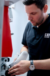 Ein Mann in schwarzem Poloshirt mit Elten-Logo richtet eine strukturierte, silbergraue Einlegesohle an einer Maschine aus.