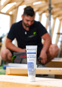 Eine Tube Physioderm UV 50 Plus steht im Vordergrund einer Holzbaustelle, im unscharfen Hintergrund arbeitet ein Handwerker.