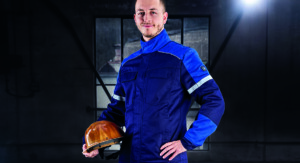 Ein Mann in dunkelblauer Schutzkleidung mit Reflexstreifen hält einen gelben Schutzhelm in der Hand und steht in einer schwach beleuchteten Industriehalle.