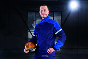 Ein Mann in dunkelblauer Schutzkleidung mit Reflexstreifen hält einen gelben Schutzhelm in der Hand und steht in einer schwach beleuchteten Industriehalle.