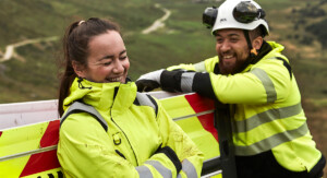 Eine Arbeiterin und ein Arbeiter in gelben Warnjacken lehnen lachend an ein Arbeitsauto vor grüner Landschaft. Der Mann trägt einen weißen Schutzhelm.