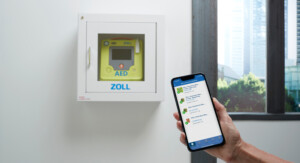 Ein automatischer externer Defibrillator in einem weißen Wandschrank der Marke Zoll ist durch das Sichtfenster sichtbar, davor hält eine Hand ein Smartphone mit der dazugehörigen App.