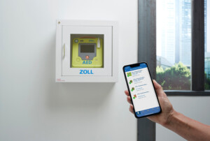 Ein automatischer externer Defibrillator in einem weißen Wandschrank der Marke Zoll ist durch das Sichtfenster sichtbar, davor hält eine Hand ein Smartphone mit der dazugehörigen App.