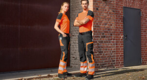 Eine Frau und ein Mann posieren vor einer Backsteinwand in orange-schwarzen Funktionsshirts und schwarz-orangen Warnschutzhosen mit breiten Reflexstreifen.