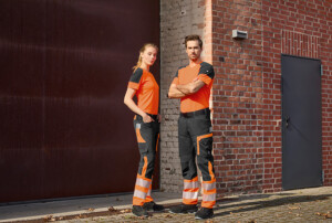 Eine Frau und ein Mann posieren vor einer Backsteinwand in orange-schwarzen Funktionsshirts und schwarz-orangen Warnschutzhosen mit breiten Reflexstreifen.