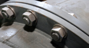 Ein Detailfoto zeigt eine Rohrflanschverbindung mit mehreren großen Sechskantschrauben und Muttern auf grauer Metalloberfläche.