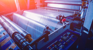 Eine Folienproduktionsanlage zeigt breite Kunststofffolie, die über mehrere Walzen läuft, daneben befindet sich ein Steuerpult mit Bedienelementen und rotem Not-Aus-Taster.