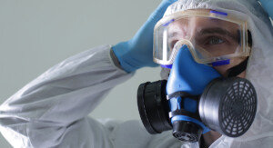 Eine Person in Schutzanzug und blauen Einweghandschuhen trägt eine Vollgesichtsatemschutzmaske mit Filtern und hält beide Hände an den Kopf.