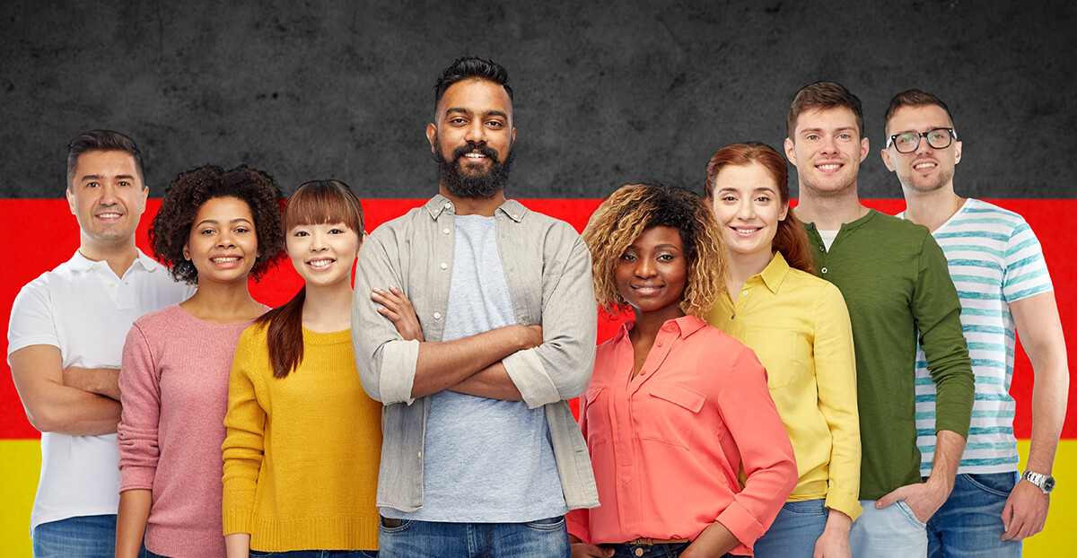 Gruppe junger Menschen unterschiedlicher Herkunft lächelt vor einer Deutschlandflagge.