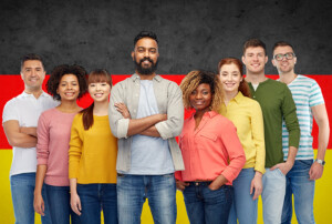 Gruppe junger Menschen unterschiedlicher Herkunft lächelt vor einer Deutschlandflagge.
