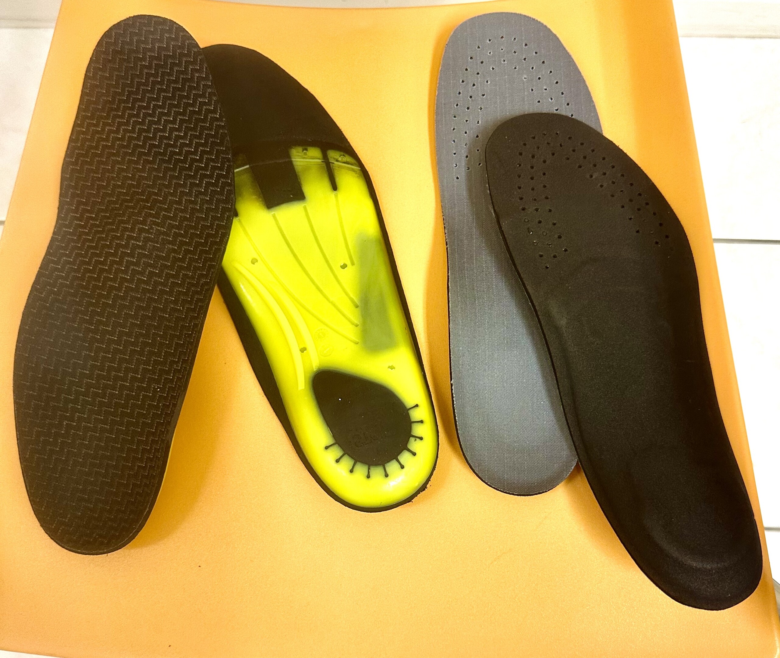 Drei verschiedene Schuheinlagen liegen auf einem orangefarbenen Stuhl; eine Einlage zeigt eine gelb-schwarze, stabile Kunststoffsohle.