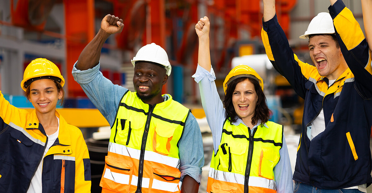 Vier Beschäftigte in Helmen und Warnwesten jubeln gemeinsam in einer Werkhalle.