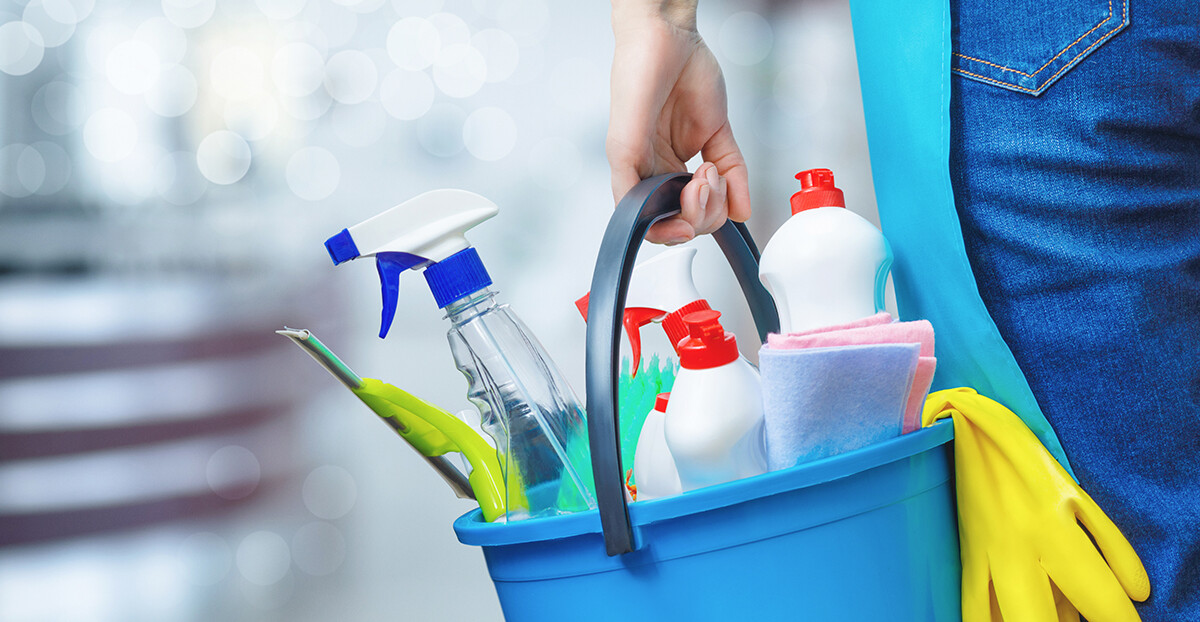 Person trägt einen blauen Putzeimer mit Sprühflasche, Reinigungsmitteln, Tuch und gelben Gummihandschuhen.