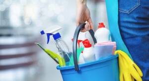 Person trägt einen blauen Putzeimer mit Sprühflasche, Reinigungsmitteln, Tuch und gelben Gummihandschuhen.