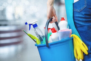 Person trägt einen blauen Putzeimer mit Sprühflasche, Reinigungsmitteln, Tuch und gelben Gummihandschuhen.
