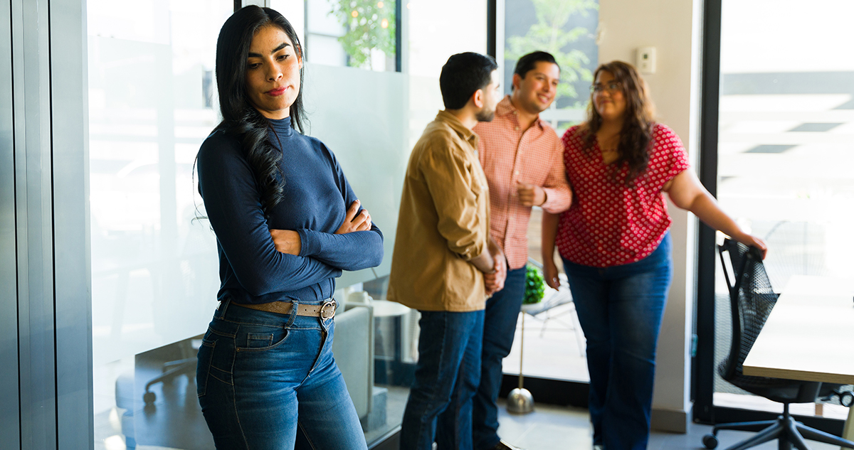 Eine Frau steht allein in einem Büro, während im Hintergrund drei Personen miteinander sprechen und lachen.
