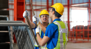 Zwei Bauarbeiter tragen Sicherheitshelme und Schutzwesten. Beide halten ein Seil und unterhalten sich. Die Szene findet auf einer Baustelle statt.