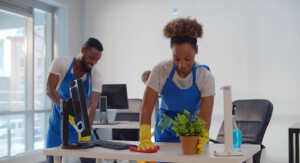Zwei Reinigungskräfte mit Handschuhen säubern Schreibtische in einem hellen Büro, Monitore und Putzmittel sind zu sehen.